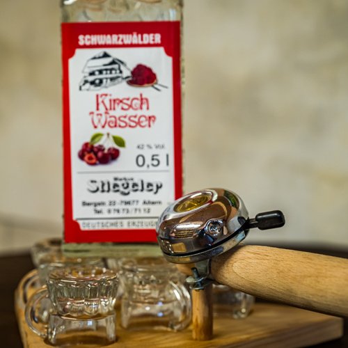 Schnapsgl&auml;ser auf einem Brett mit Radklingel im Hintergrund eine Flasche Kirschwasser  - 